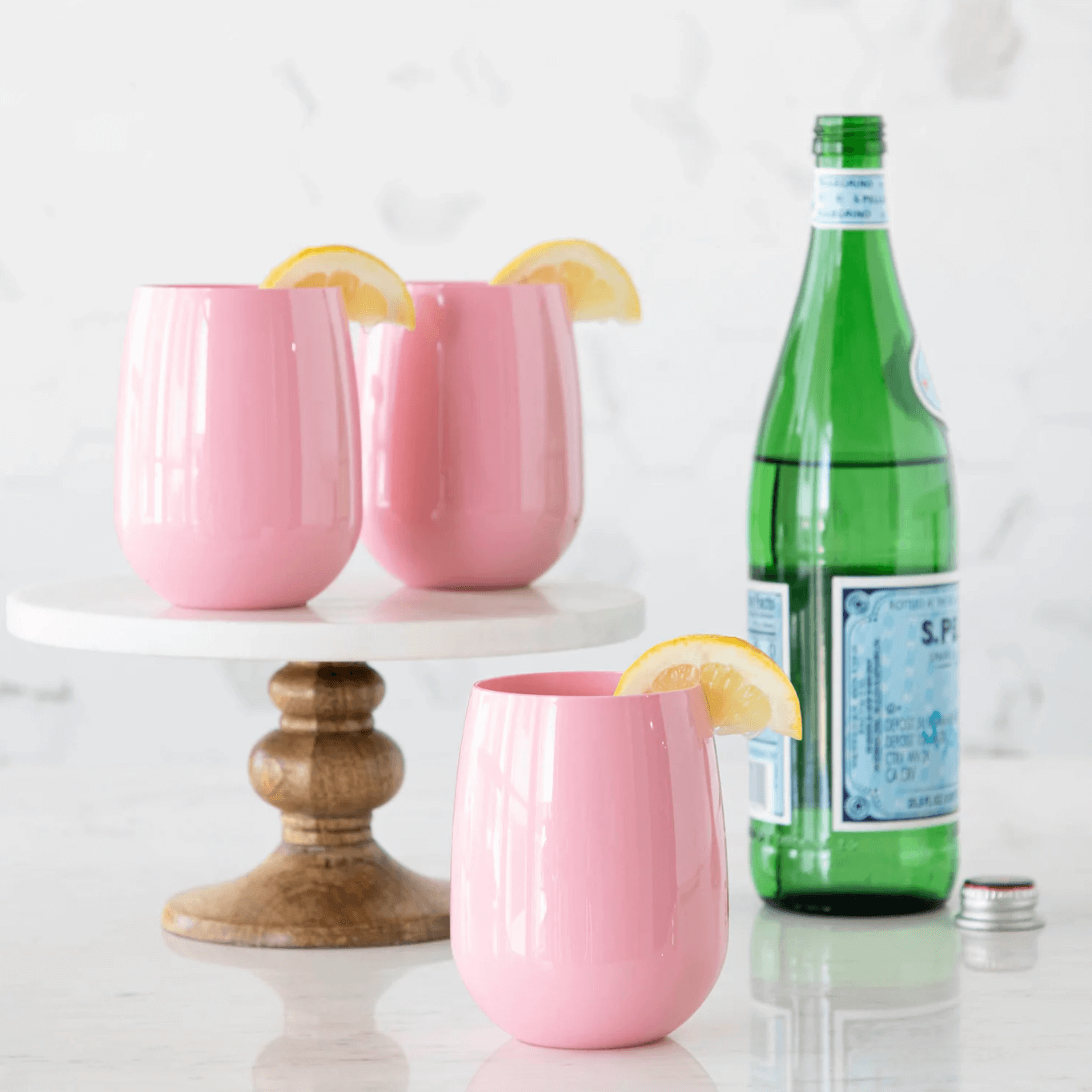 Three pink cups with lemon wedges on a cake stand, alongside a fourth cup and a green glass bottle of sparkling water on a marble surface—perfect for serving refreshing summer gifts.