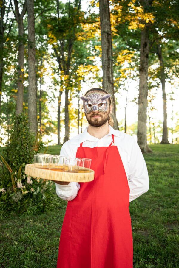 A Wild Storybook Wedding You Won't Believe - 17 Server in a mask holding a tray of whiskey shots in the woods