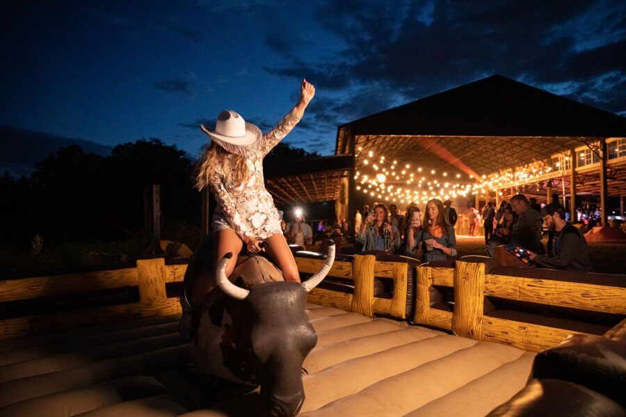 A Wild Storybook Wedding You Won't Believe - 7 Kimberlin bride rides a mechanical bull with the Friday welcome part barn in the background