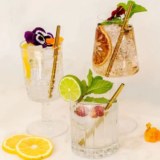 Three glasses of cocktails with garnishes including flowers, lemon slices, a dried orange slice, lime, raspberries, and mint leaves. Gold straws are in each glass.