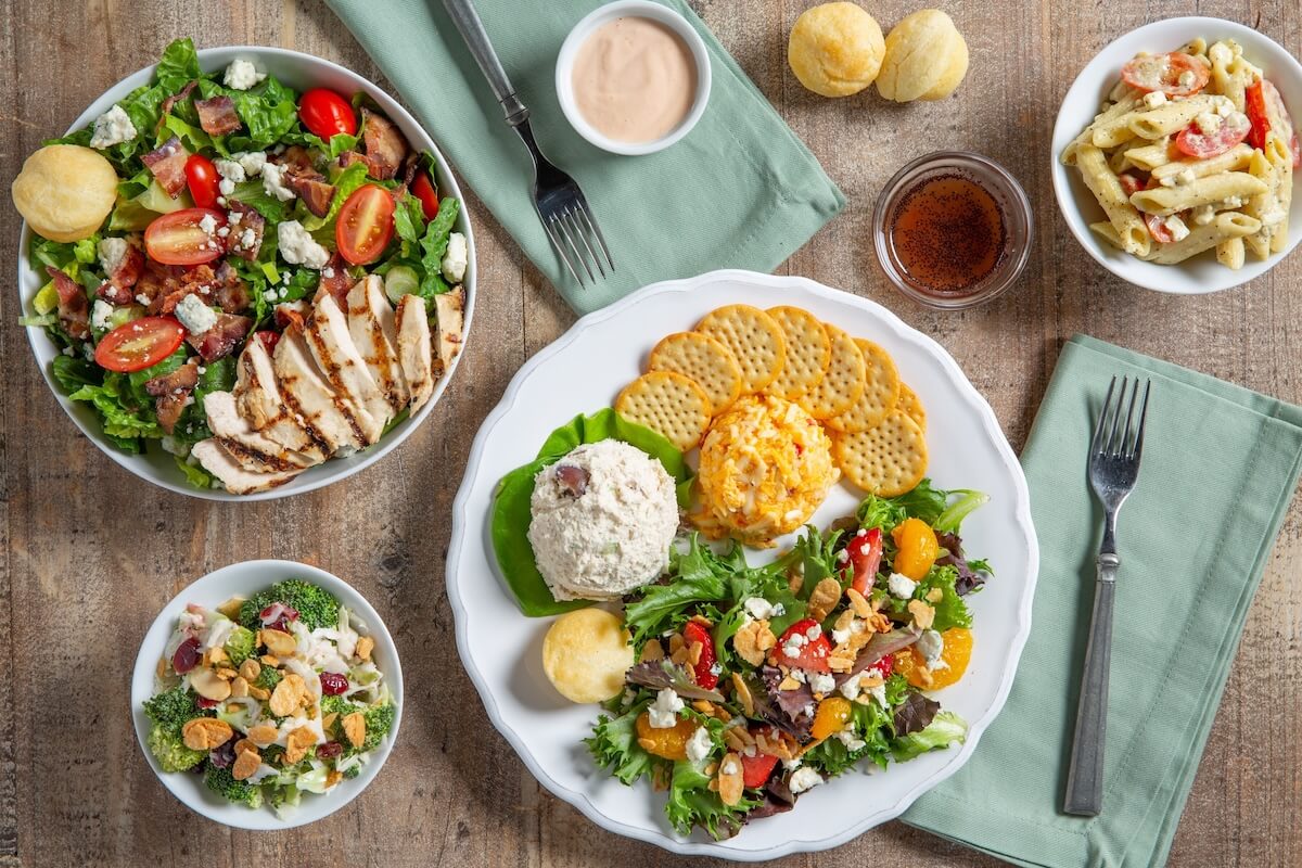 An overhead view of a table in Cahaba Heights set with salads, crackers, chicken salad, pasta salad, cheese spread, rolls, dressing, and two forks on green napkins.