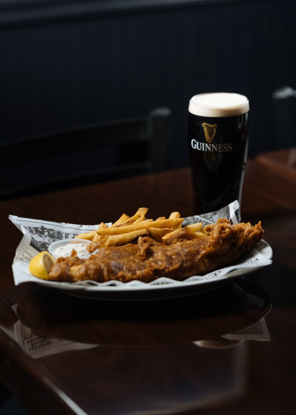 Plate of fish and chips with a lemon wedge, served alongside a pint of Guinness on a dark wooden table, perfect for group dining in Nashville.