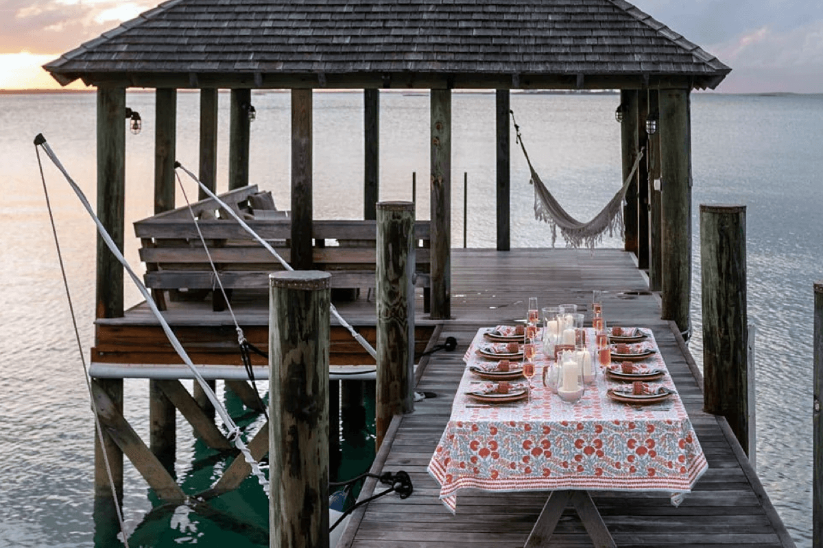A wooden dock over water with a table set for a meal, featuring plates, glasses, and candles on a patterned tablecloth. A hammock finds its place between two posts near the table, and a covered structure is visible in the background.