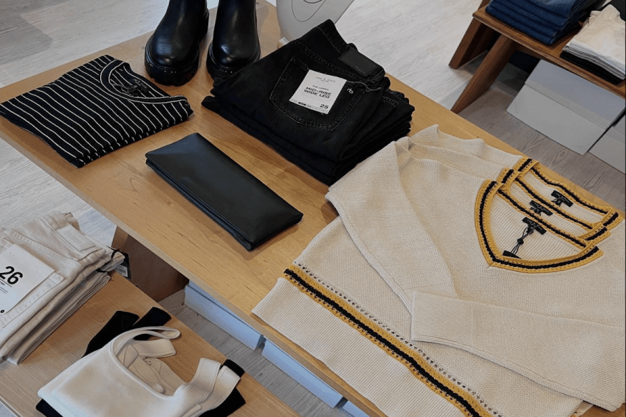 Discover the latest styles at Louisville boutiques with this clothing display featuring a striped shirt, black jeans, a cream sweater, black boots, a black wallet, folded pants, and a white crop top on a wooden table.