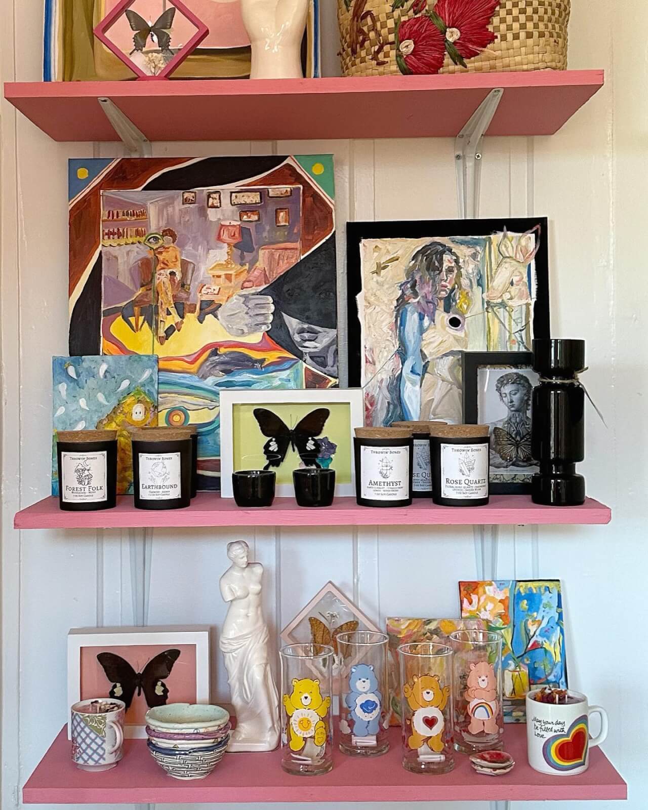 three pink wooden shelves filled with artwork, candles, glasses, and more