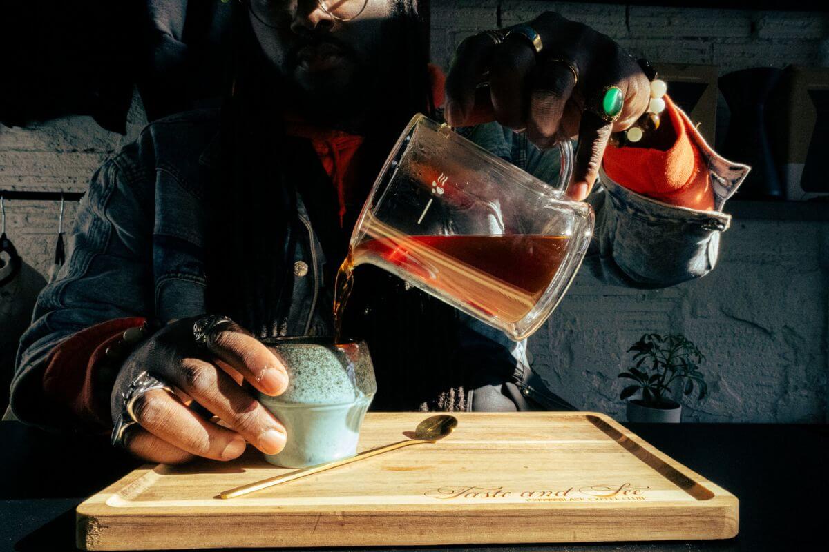 Person pouring coffee from a glass carafe into a cup on a wooden tray, with “Taste and See” engraved on it—a cozy moment inspired by the charm of Memphis coffee shops.