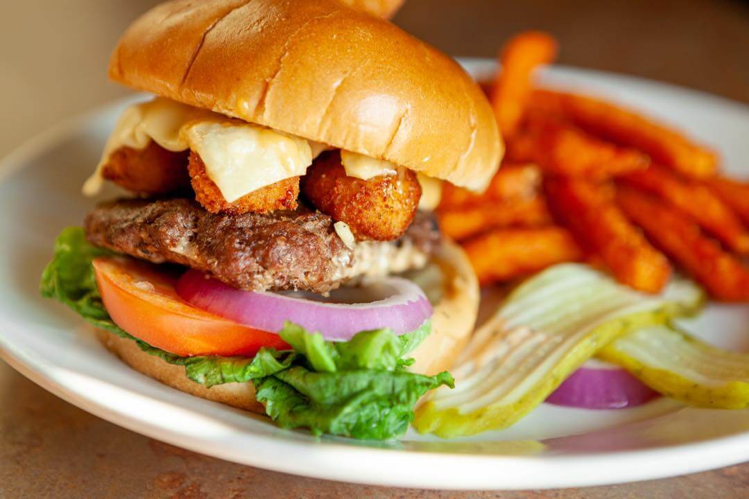 A close-up of a burger with lettuce, tomato, onions, cheese, and fried toppings on a bun, served with a side of sweet potato fries and pickles—a perfect meal to enjoy while catching the University of Missouri football game.