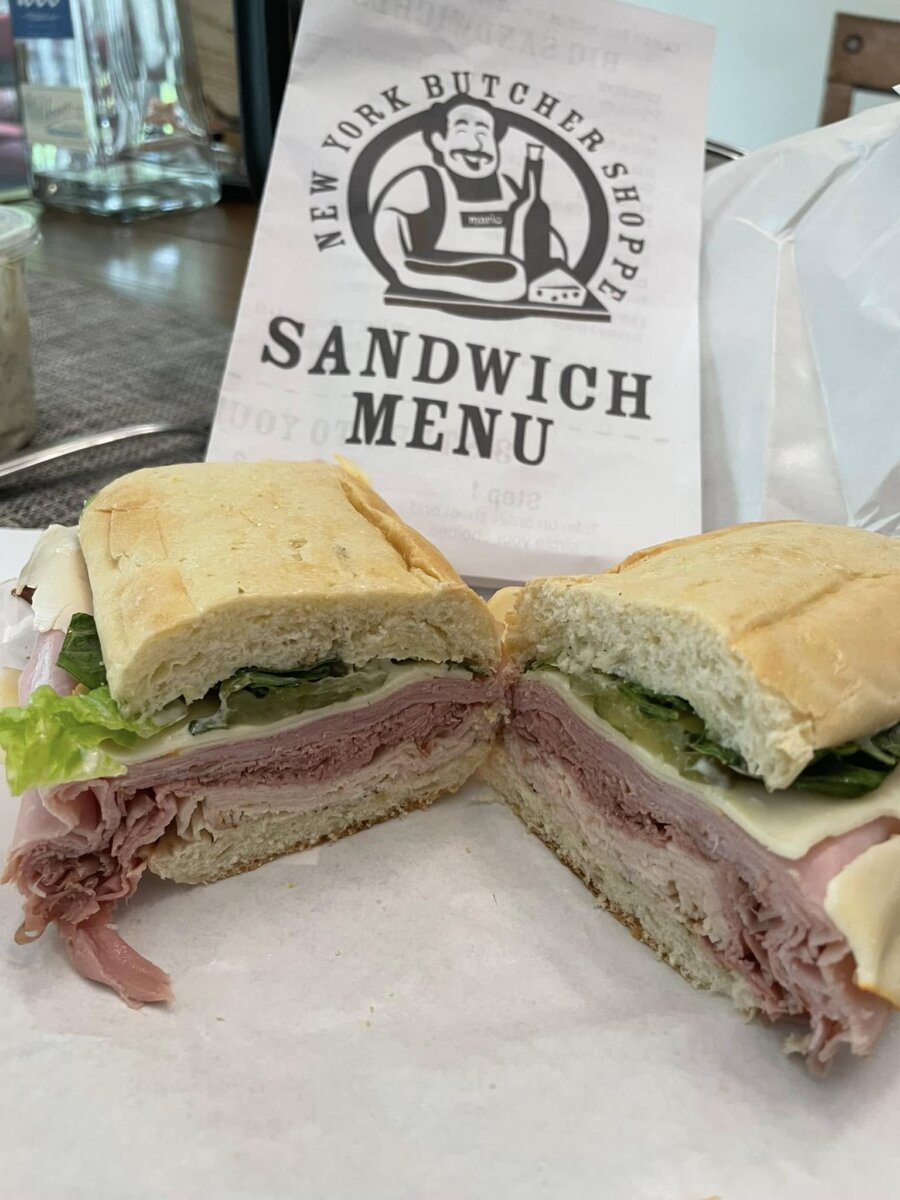 A ham and lettuce sandwich on a baguette, considered one of the best lunch sandwiches, is sliced in half and displayed in front of a New York Butcher Shoppe sandwich menu.