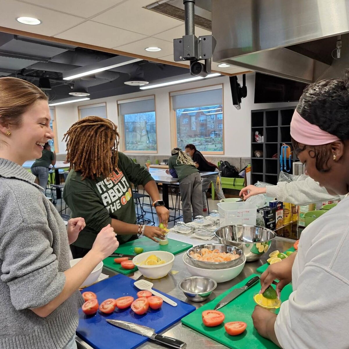 8 Ways to Meet New People in BHAM - 5 Three people prepare vegetables together at a kitchen counter, slicing tomatoes and cucumbers—a great example of how to meet people in Birmingham through shared cooking experiences—while others sit at tables in the background.