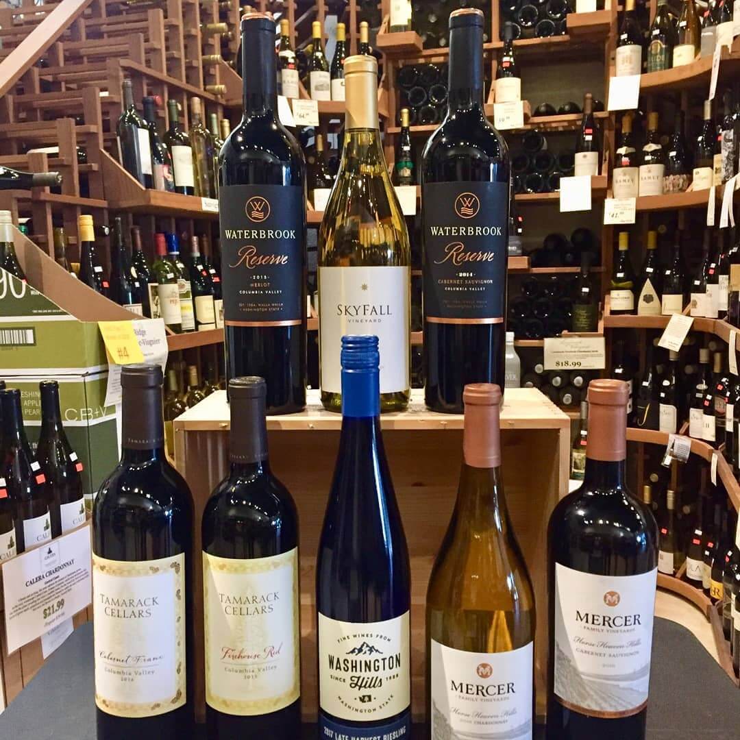 Eight wine bottles are lined up front and center with The Vintage Wine Shoppe's vast wine selection in the background. 