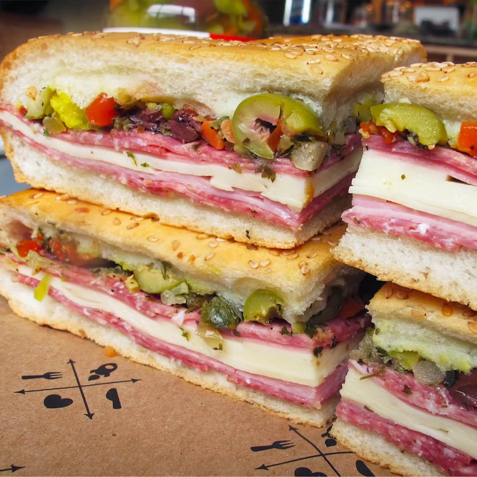 Four stacked slices of muffuletta sandwich with layers of meats, cheese, and olive salad on sesame bread—just like those from southern restaurants that ship—placed on a cardboard surface.