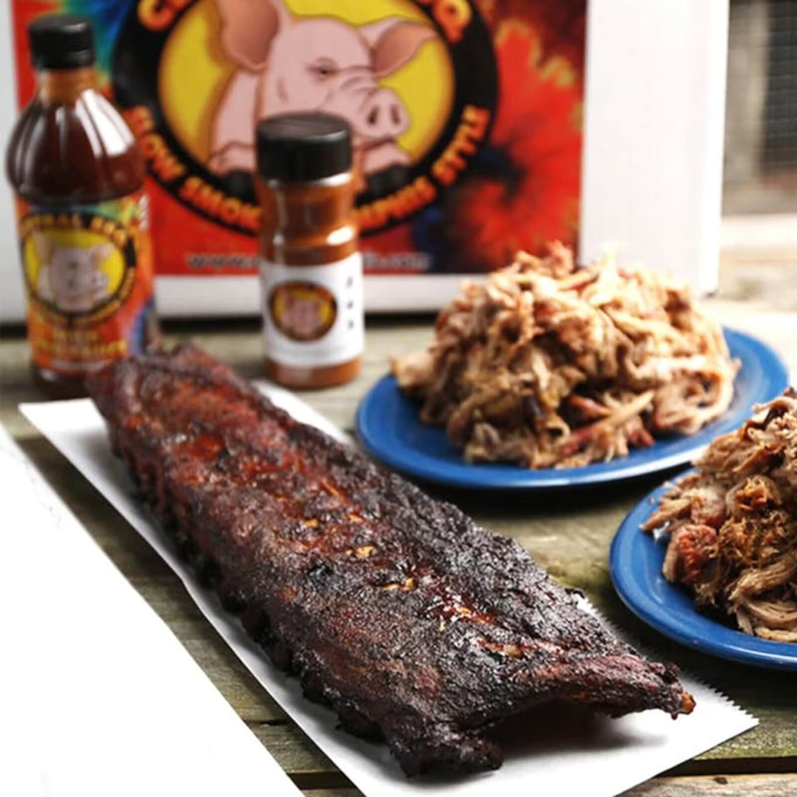 A slab of barbecued ribs on a tray, with plates of pulled pork and bottles of barbecue sauce and seasoning in the background—just like the flavors from southern restaurants that ship straight to your door.