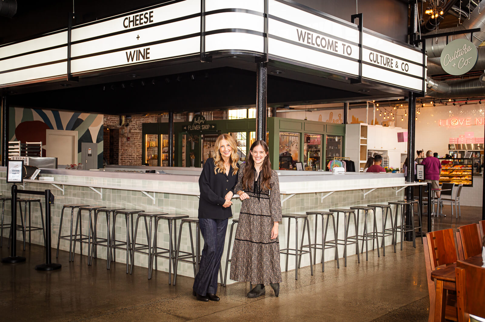 Two women in front of a cheese and wine bar with stools