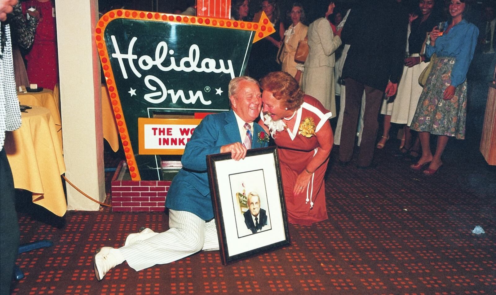 Kemmons and Dorothy pose with a portrait in front of a Holiday Inn Sign