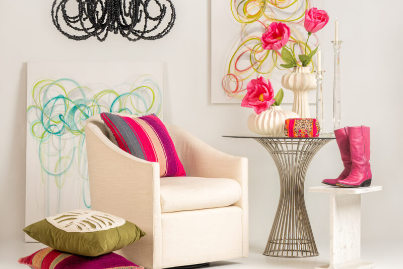 A cozy living space featuring an armchair with colorful cushions, a glass-top side table with floral decor, a pink boot from the best gift shops in Memphis, abstract wall art, and a modern chandelier.