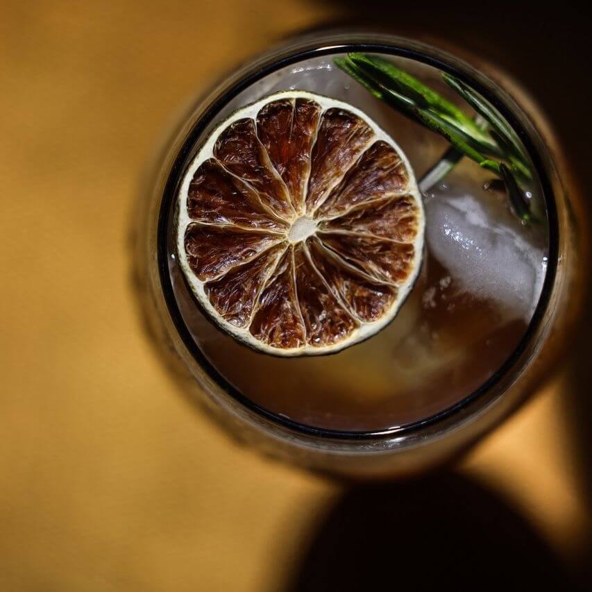 Your Updated Guide to Crosstown - 2 A close-up of a drink with ice, garnished with a dried citrus slice and a green leaf, photographed from above on a yellow surface at Crosstown Concourse.