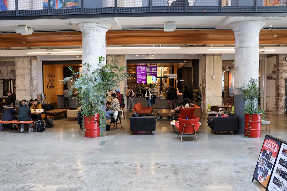 People sit and socialize in a modern, open cafe space at Crosstown Concourse, surrounded by concrete columns, red planters, and a counter in the background.
