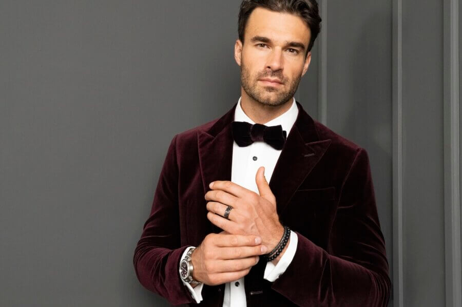 Discover Nashville’s Luxury Secret - 5 Man in a burgundy velvet tuxedo jacket with a black bow tie poses against a gray wall, accessorized with bracelets and a watch from Diamond Cellar.
