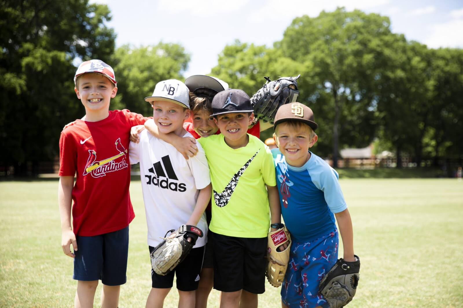 Five young boys wearing baseball gear stand together on a grassy field with trees in the background, embodying the spirit of Nashville summer camps.