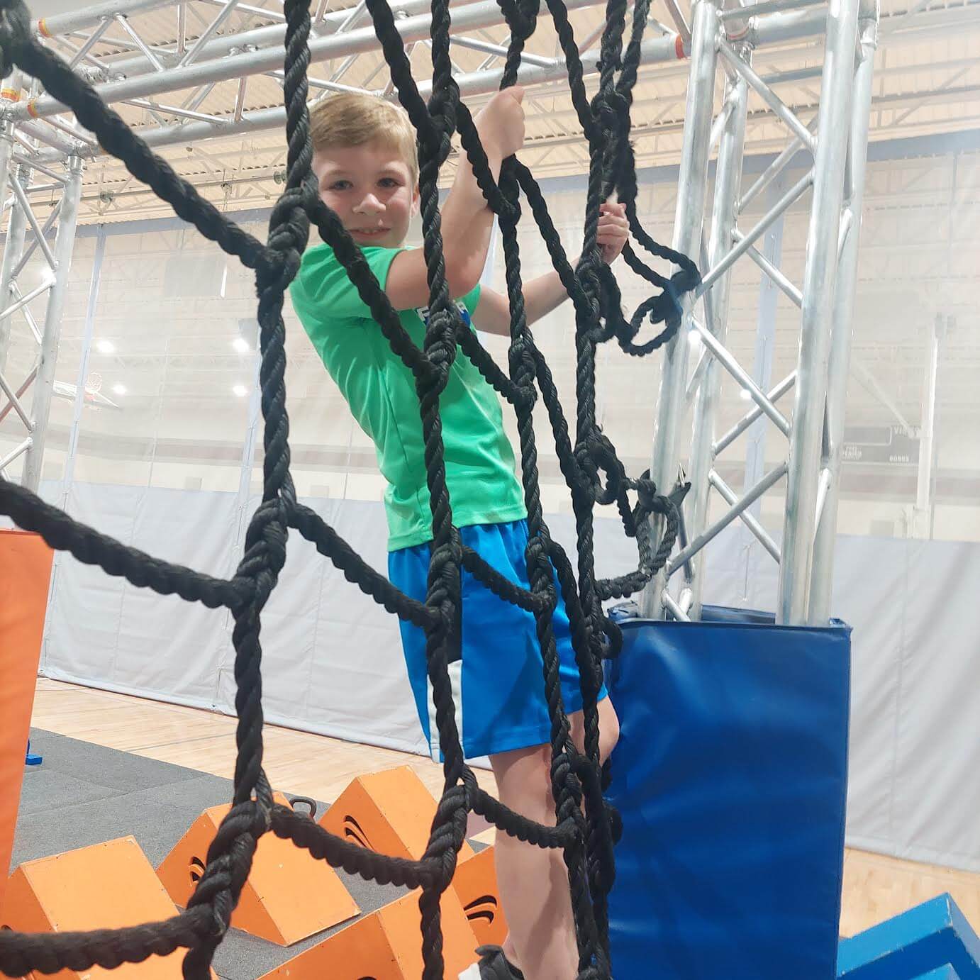 A child in a green shirt and blue shorts climbs a rope net in an indoor obstacle course, reminiscent of those found at Memphis summer camps. Orange foam blocks are visible below, adding a splash of color to the adventurous scene.