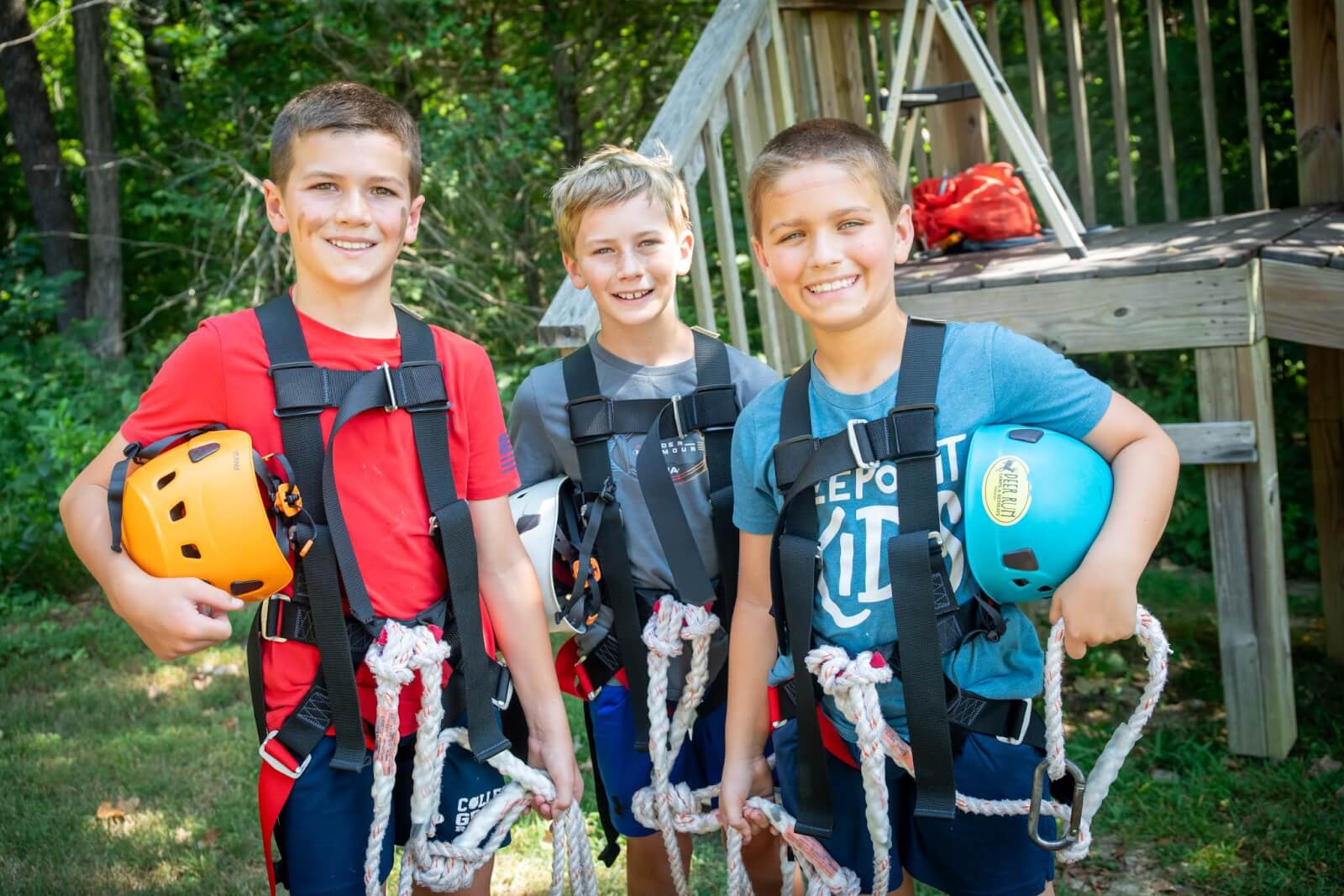 Three boys in harnesses and helmets stand smiling near a wooden structure, holding ropes and safety gear, ready for adventure. They’re experiencing the thrill of Nashville summer camps, where every day is filled with excitement and outdoor fun.