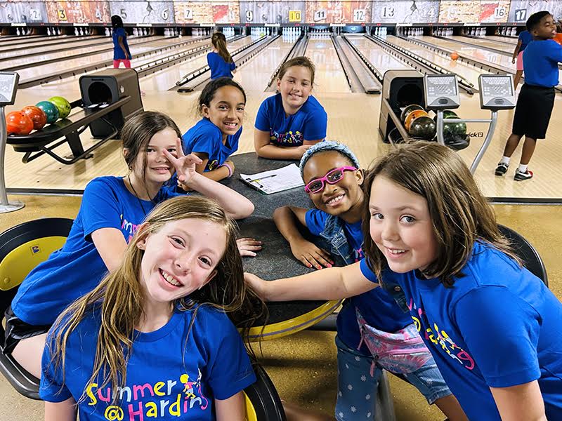 A group of six children in blue shirts, fresh from their Memphis summer camps, are all smiles as they pose at a bowling alley. They sit around a table with vibrant bowling lanes and colorful balls adding to the lively atmosphere in the background.