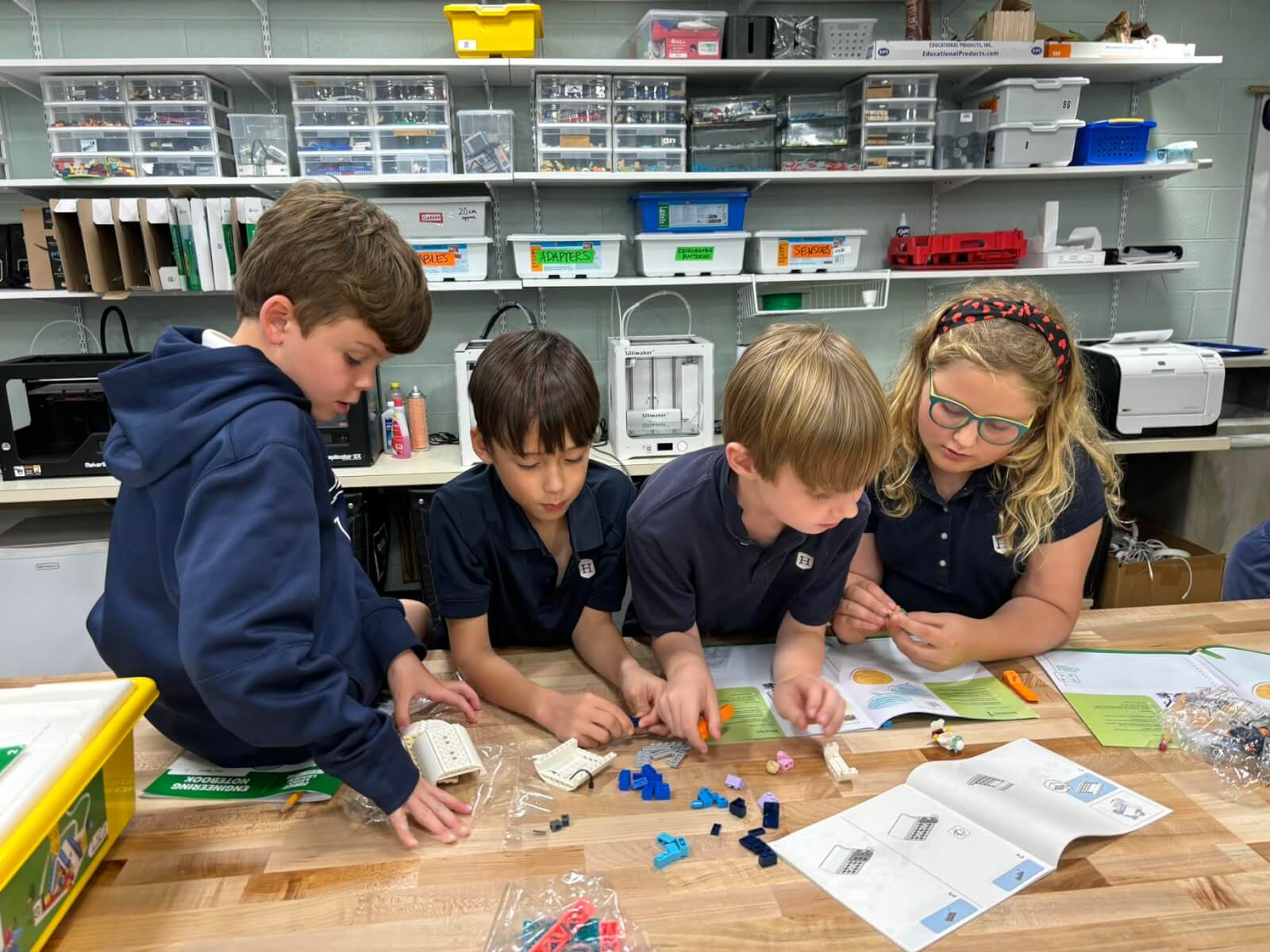 In a Nashville summer camp, four children in a classroom collaborate on a building project, carefully following instructions and assembling small parts. Shelves brimming with supplies are visible in the background, fueling their creativity and teamwork.