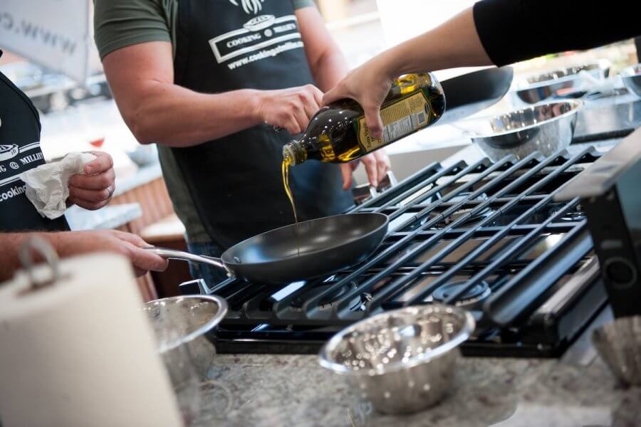 An instructor pours oil into a frying pan on the stove, surrounded by eager participants in cooking aprons, during one of Louisville's engaging adult education classes.