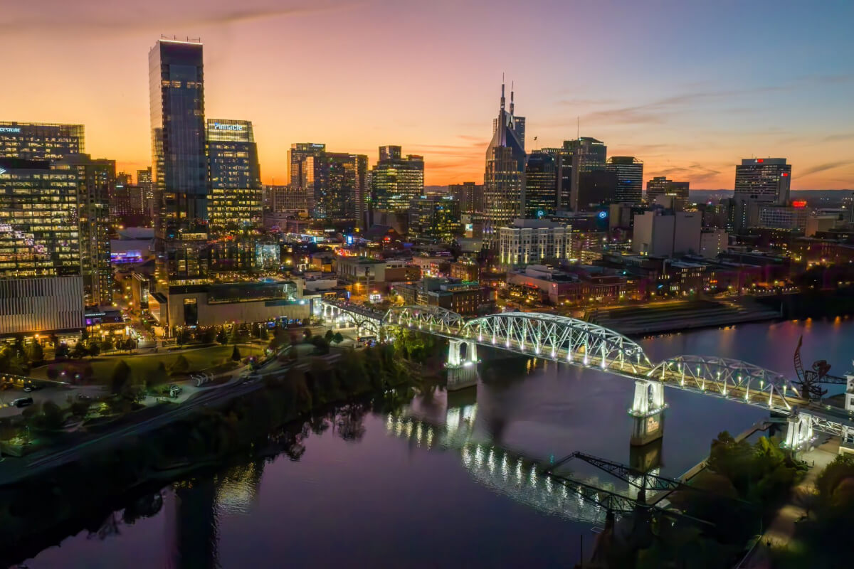 A cityscape at sunset showcases a lit-up bridge over a river, with illuminated skyscrapers and a colorful sky in the background, reminiscent of the vibrant atmosphere during Nashville events in February.