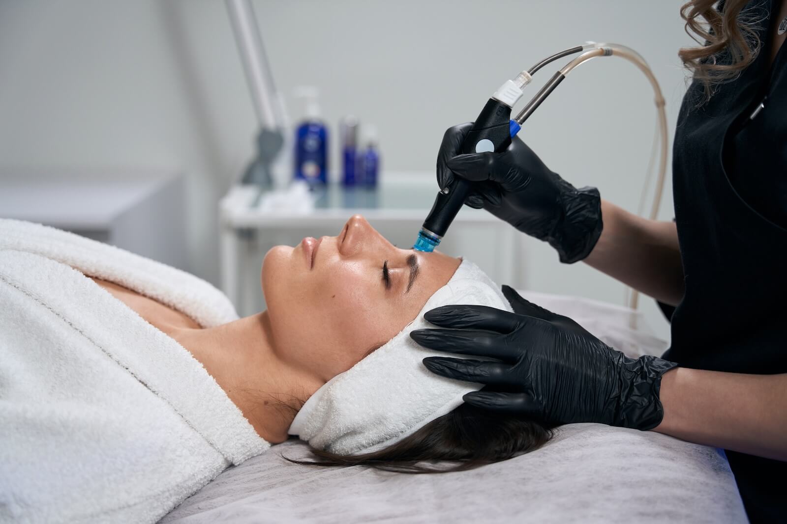 Cosmetologist in protective gloves sitting near beautiful lady and doing HydraFacial procedure in salon