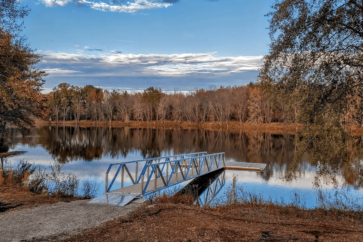 A small metal dock extends into a calm lake surrounded by trees with autumn foliage, under a partly cloudy blue sky—much like the tranquility found at Memphis events in February.