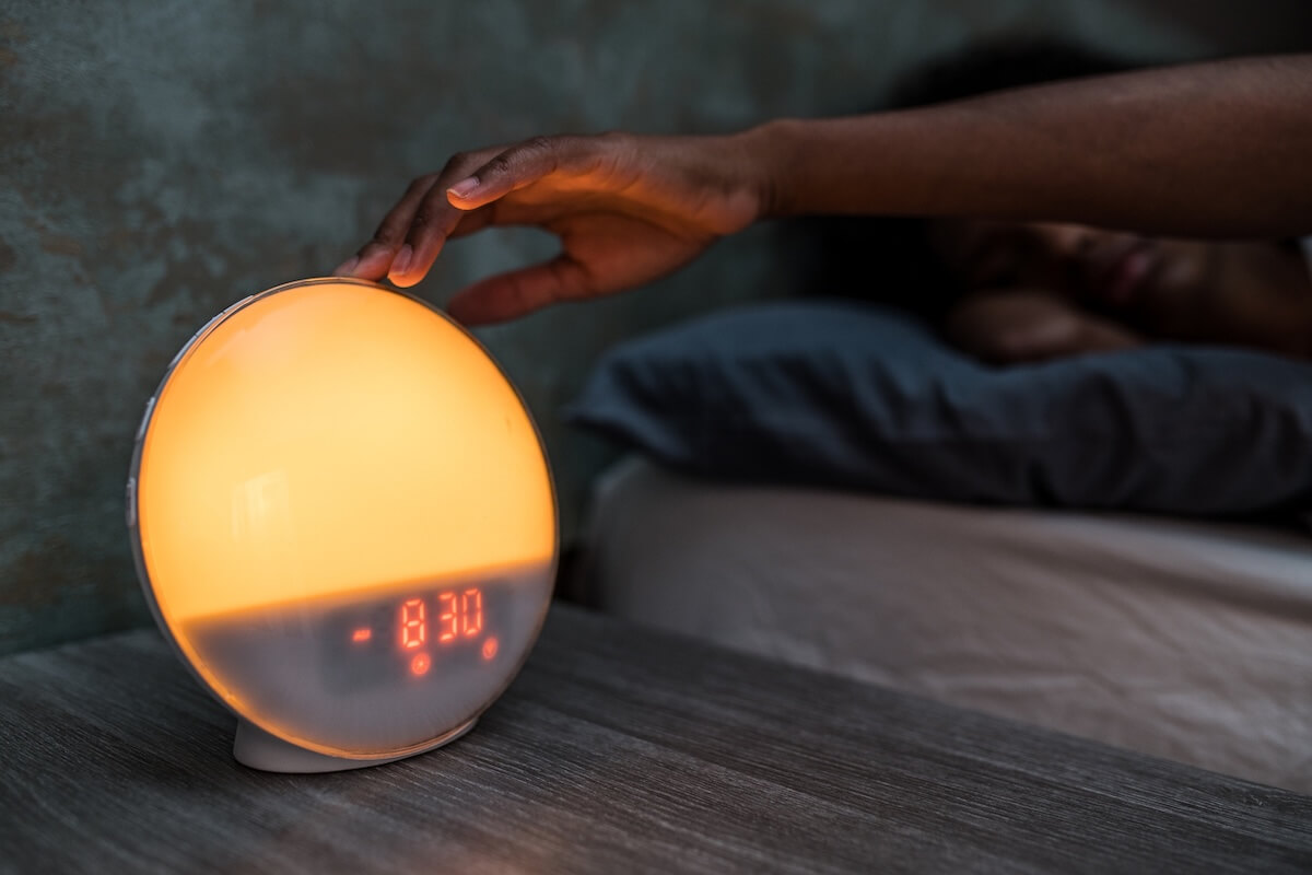 A hand reaches for an illuminated round alarm clock on a bedside table, as the gentle whisper of cortisol ensures the person sleeping in the background starts their day refreshed and ready.
