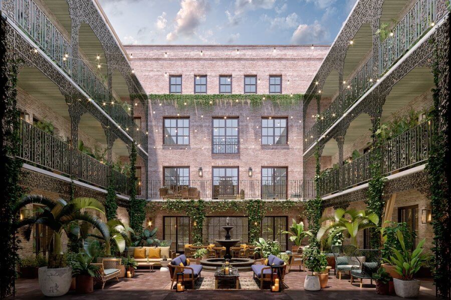 23 New Boutique Hotels in the South - 16 A courtyard in new boutique hotels features multiple stories of brick buildings, iron railings, and lush greenery. Lounge seating encircles a central fountain while string lights dangle above under a partly cloudy sky.