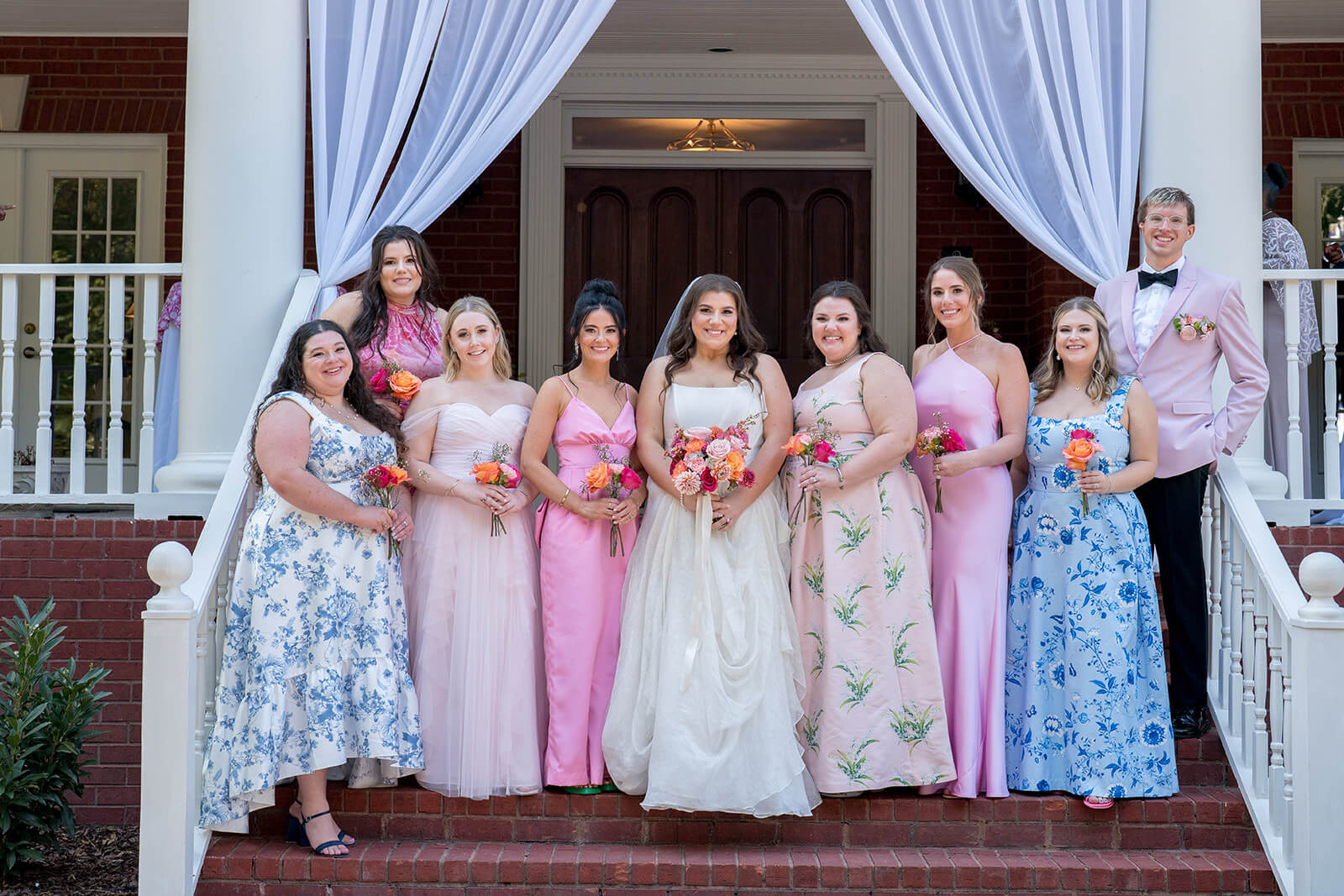 The Top Bridal Boutiques in Nashville - 3 A bride and nine bridesmaids in pink and floral dresses pose on brick steps of a white house with draped curtains, showcasing elegance that rival the top bridal boutiques in Nashville.