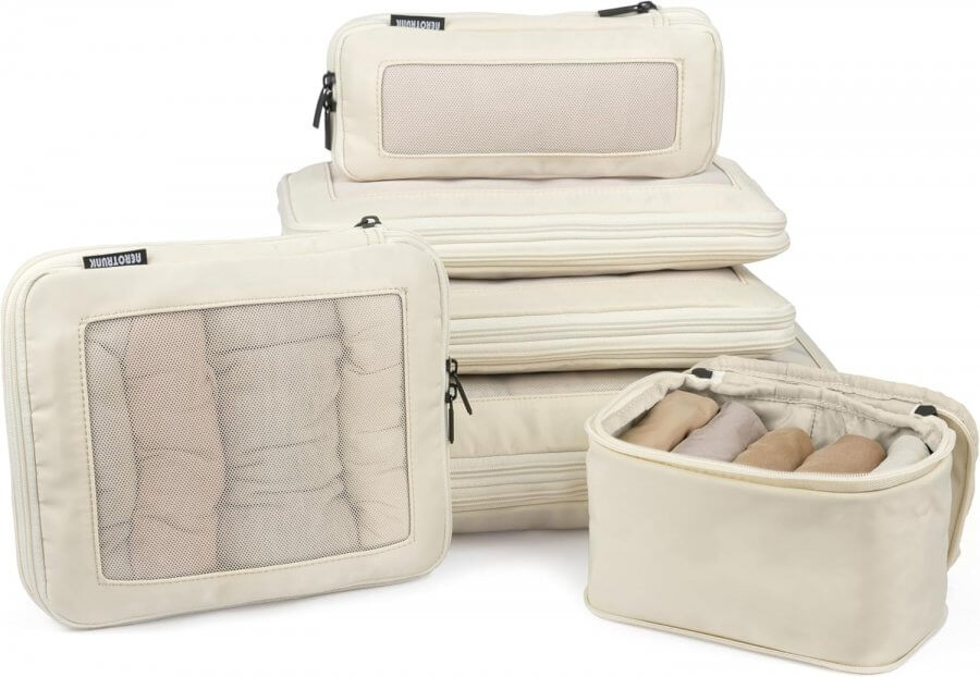 A set of beige packing cubes, considered among the best travel accessories, filled with neatly rolled clothes. The set includes various sizes, with some featuring mesh panels and zippers for easy access.