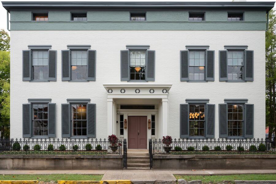 23 New Boutique Hotels in the South - 11 A two-story white brick house with green shutters and a central door framed by columns stands gracefully behind a sleek black iron fence.