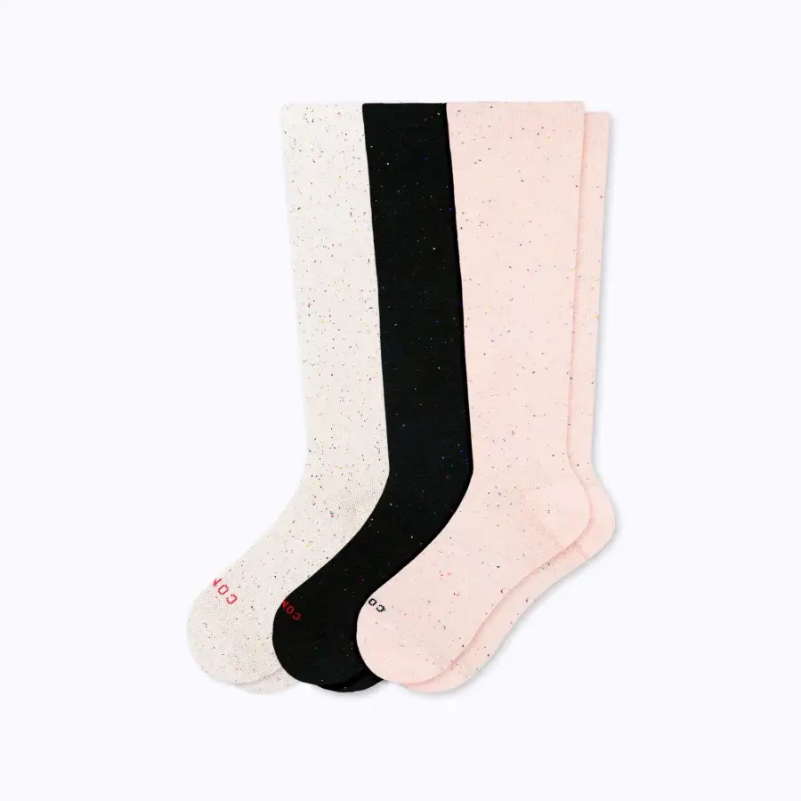 Three knee-high socks are laid out flat: one off-white, one black, and one pink, all with a speckled pattern. These cozy essentials make for the best travel accessories, ensuring comfort and style on every journey.