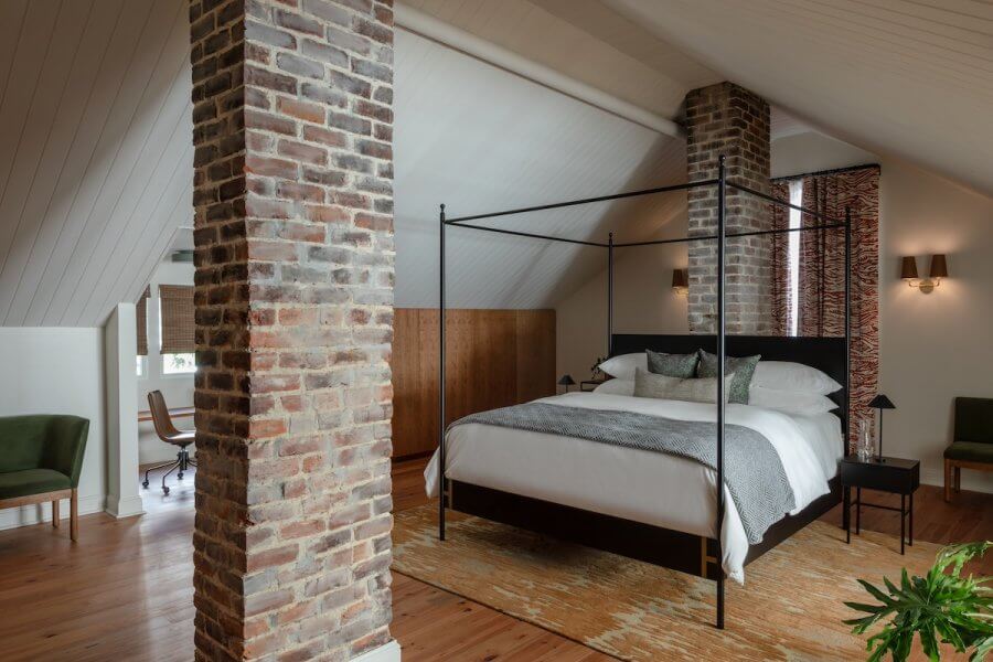 23 New Boutique Hotels in the South - 4 Cozy bedroom in one of the new boutique hotels, featuring a canopy bed, brick columns, wood flooring, an office desk, and green chairs. Neutral and earthy tones elegantly complement the space.