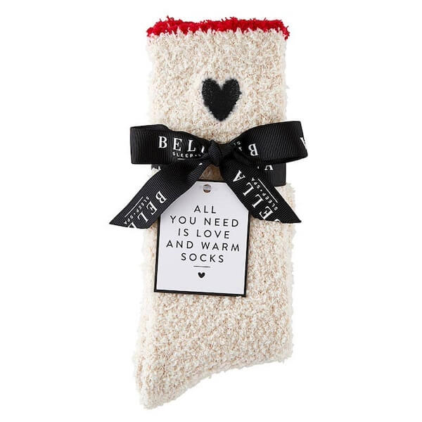 Fluffy beige socks featuring a black heart design, tied elegantly with a black ribbon. A tag reads, "All you need is love and warm socks." Discover this cozy delight in Southern FINDS this February.