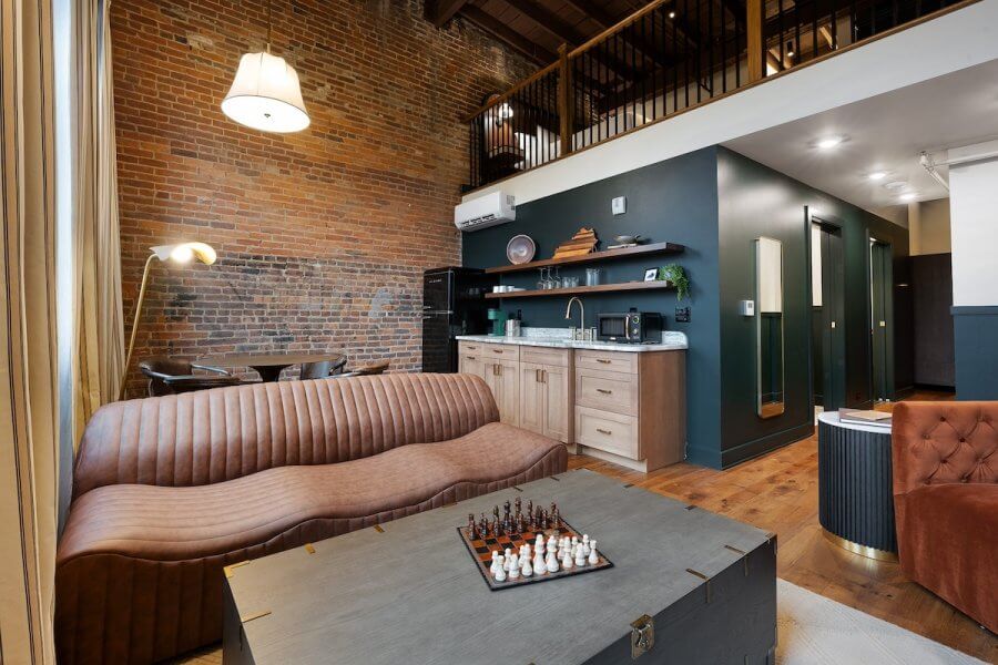 23 New Boutique Hotels in the South - 8 Cozy living space with brick walls, a brown sofa, and a chess set on the table, reminiscent of new boutique hotels. A kitchenette with modern appliances and a loft area above are visible. Warm lighting creates an inviting atmosphere.