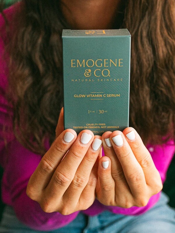 Against a purple background, a person with painted nails proudly displays a green box of Emogene & Co. Glow Vitamin C Serum. This Southern FINDS favorite brings February freshness to your skincare routine with its vibrant boost of radiance.