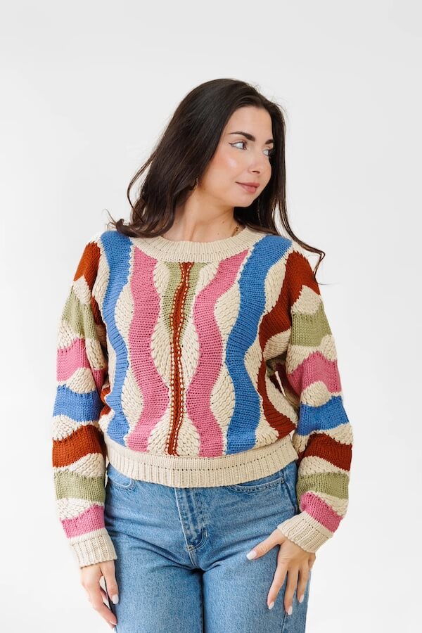 A person in a colorful, wavy-patterned sweater reminiscent of Southern charm, with shades of pink, blue, brown, and green, paired with blue jeans. Set against a plain white backdrop, this February-inspired ensemble captures the essence of cozy style.