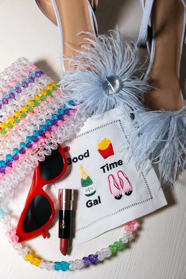 A pair of shoes with blue feathers, a colorful beaded bag, and red sunglasses lay next to lipstick and a napkin emblazoned with "Good Time Gal" alongside sketches of wine, fries, and shoes—perfect for your February Southern FINDS.