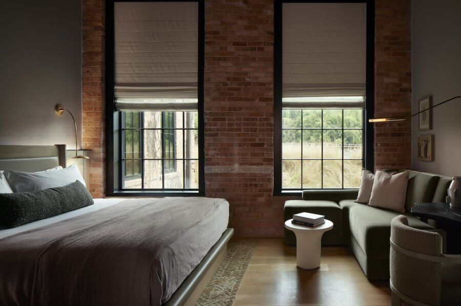 23 New Boutique Hotels in the South - 2 A cozy bedroom reminiscent of new boutique hotels features brick walls, a large bed, a sofa by the window, and a small round side table. Two large windows with Roman shades let in abundant natural light.