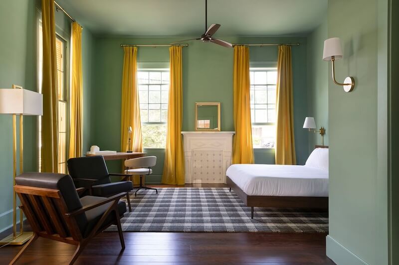 23 New Boutique Hotels in the South - 6 The bedroom, reminiscent of new boutique hotels, features green walls, large windows with yellow curtains, a white bed, wooden furniture, a checkered rug, and a ceiling fan.