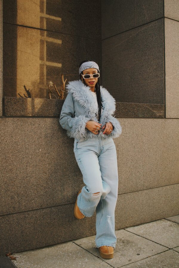 A woman in jeans and a furry jacket leaning against a wall.