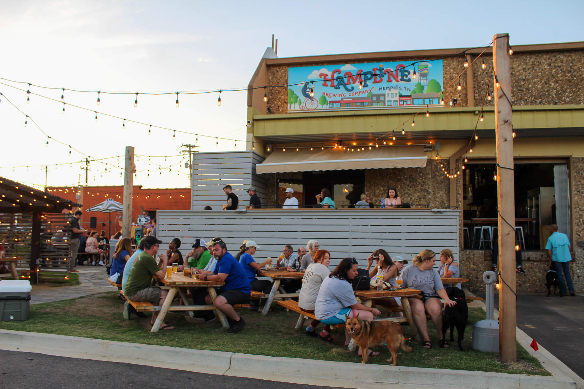 People sit at picnic tables outside a restaurant with string lights overhead; some have dogs with them, and a colorful sign for “Tampine” is visible on the building, creating a lively spot perfect for Memphis trivia nights.