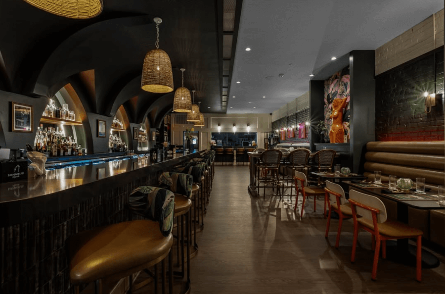 23 New Boutique Hotels in the South - 10 A dimly lit, modern bar nestled within one of the new boutique hotels features an arched ceiling, wooden bar stools, wicker pendant lights, and a row of tables. The ambiance is warm, with contemporary artwork adorning the walls.