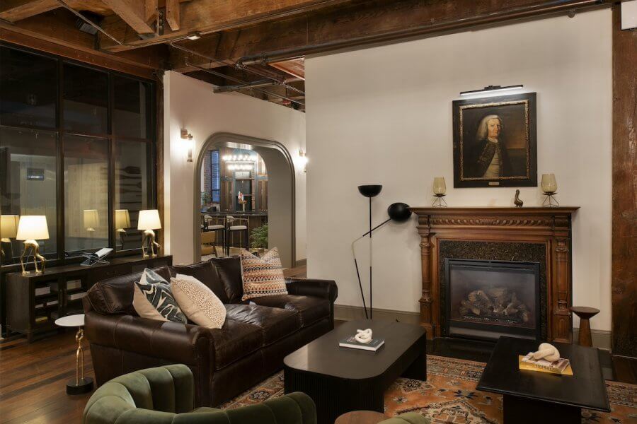 23 New Boutique Hotels in the South - 9 Stylish living room featuring a brown leather sofa, decorative cushions, and a dark wood coffee table. A painting hangs above the ornate fireplace, while warm lighting and exposed wood beams enhance the cozy ambiance.