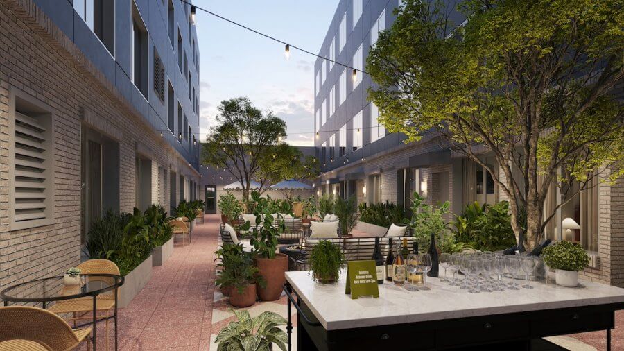 23 New Boutique Hotels in the South - 17 The courtyard of the new boutique hotel features patio furniture, string lights, and potted plants lining a pathway. A table holds wine bottles, glasses, and a small sign. Nearby, multi-story buildings with windows create an inviting backdrop.